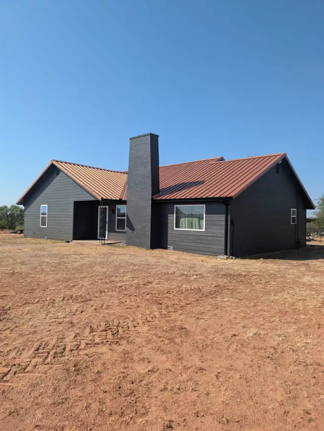 Dark grey steel home with a rust-red roof and a brick chimney, seen from the front on red dirt