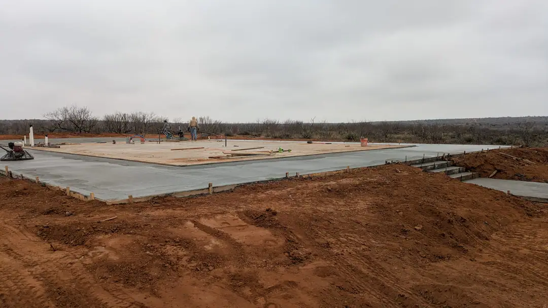 Crew finishing a fresh concrete slab on red dirt under an overcast sky