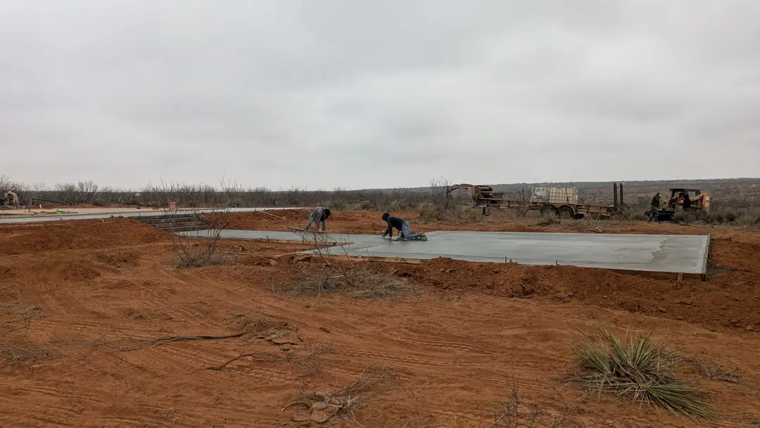 Two crew members hand-finishing the edge of a new slab with a skid steer and trailer behind