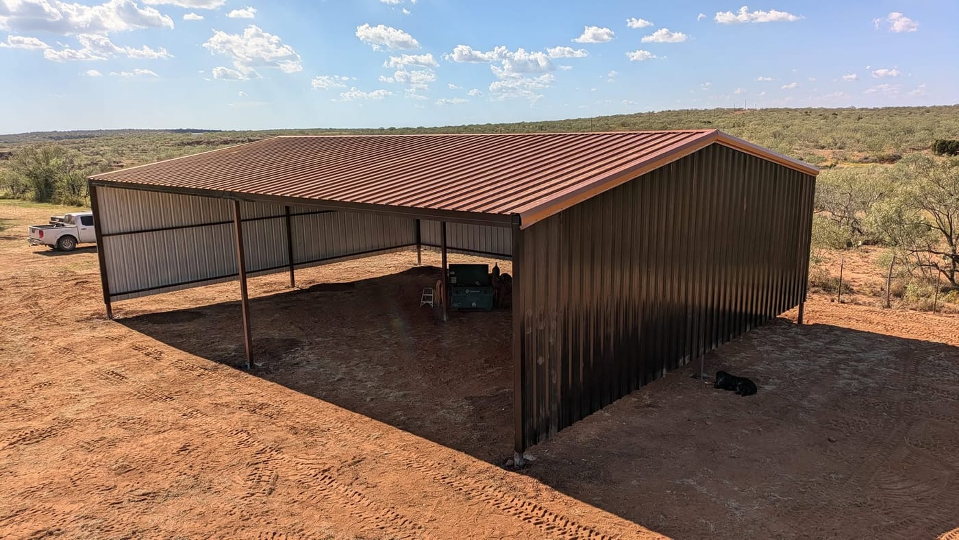 Open-sided steel shed with a brown roof and dark walls on the ranch