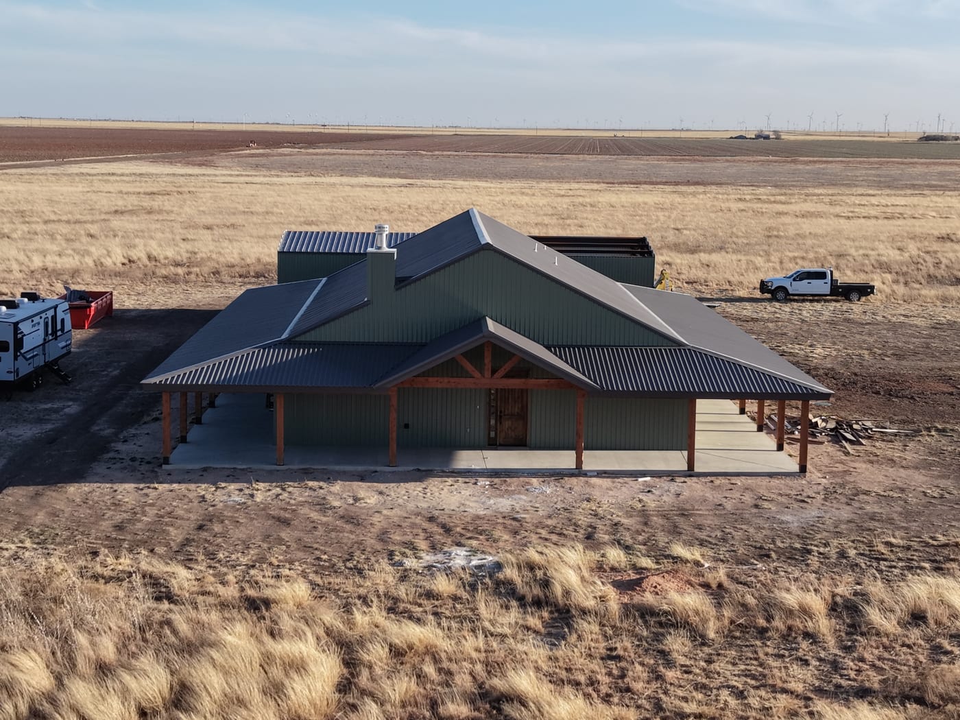 Drone view of a sage green steel home with a timber-framed front porch on open farmland