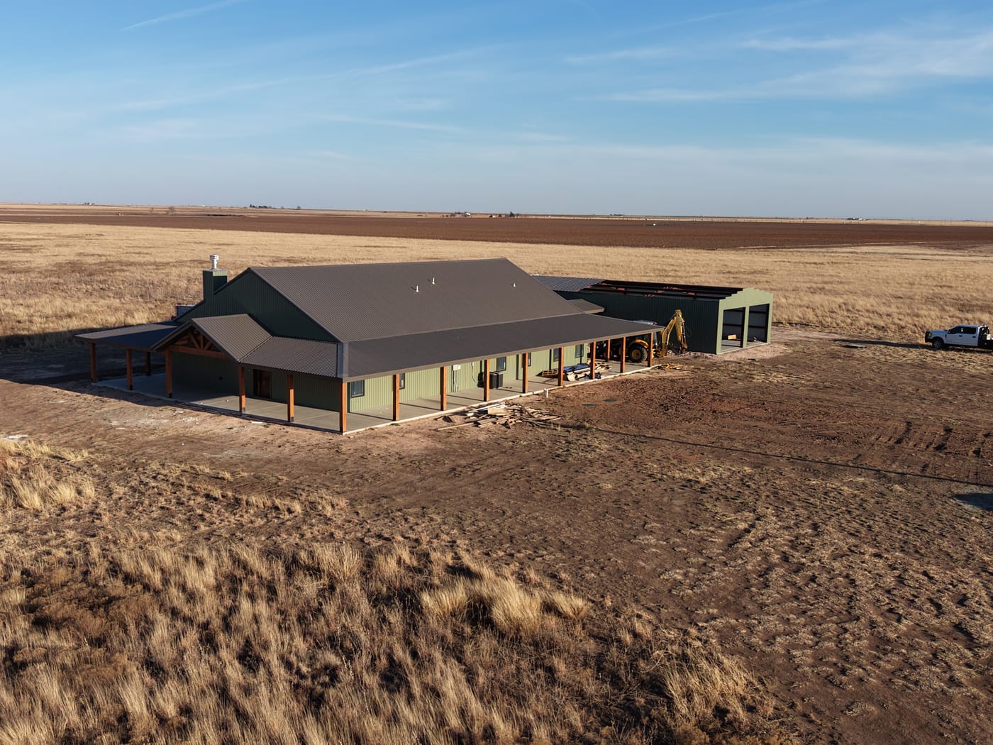 Drone view of a sage green steel home with a long covered porch and an attached shop going up
