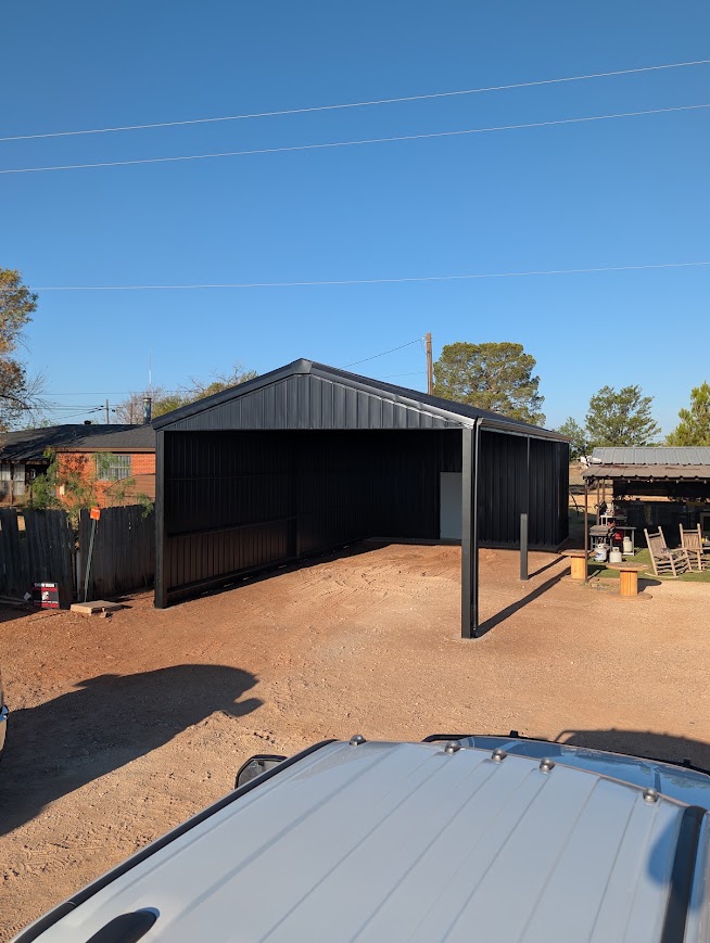 Black open-front steel barn beside a backyard patio, seen from a truck