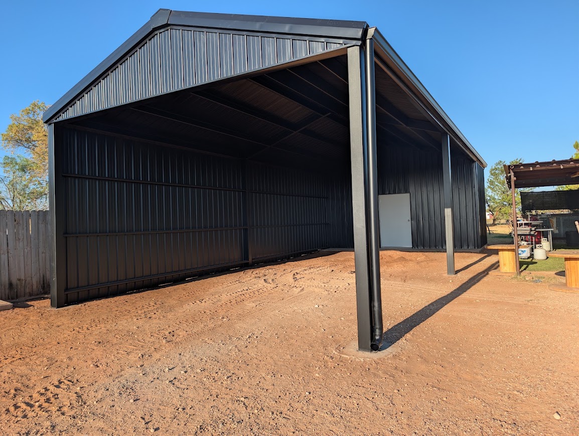 Black open-front steel barn with a white walk door on red dirt under a blue sky